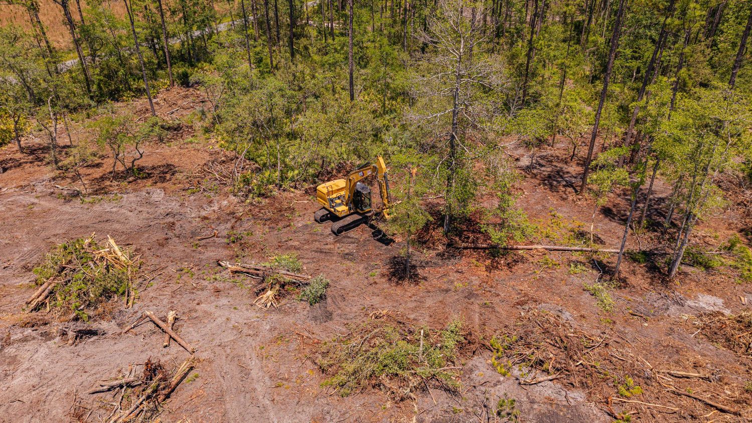 Aerial view of a yellow bulldozer clearing forest land, surrounded by felled trees and red clay soil.