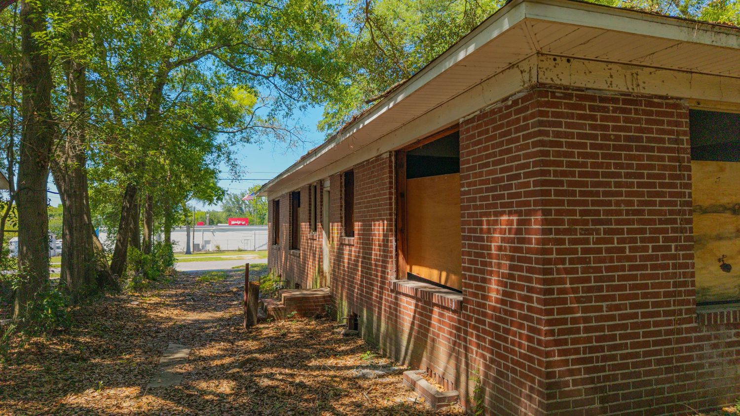 Abandoned brick building with boarded windows surrounded by tall trees and overgrown grounds in a rural setting.