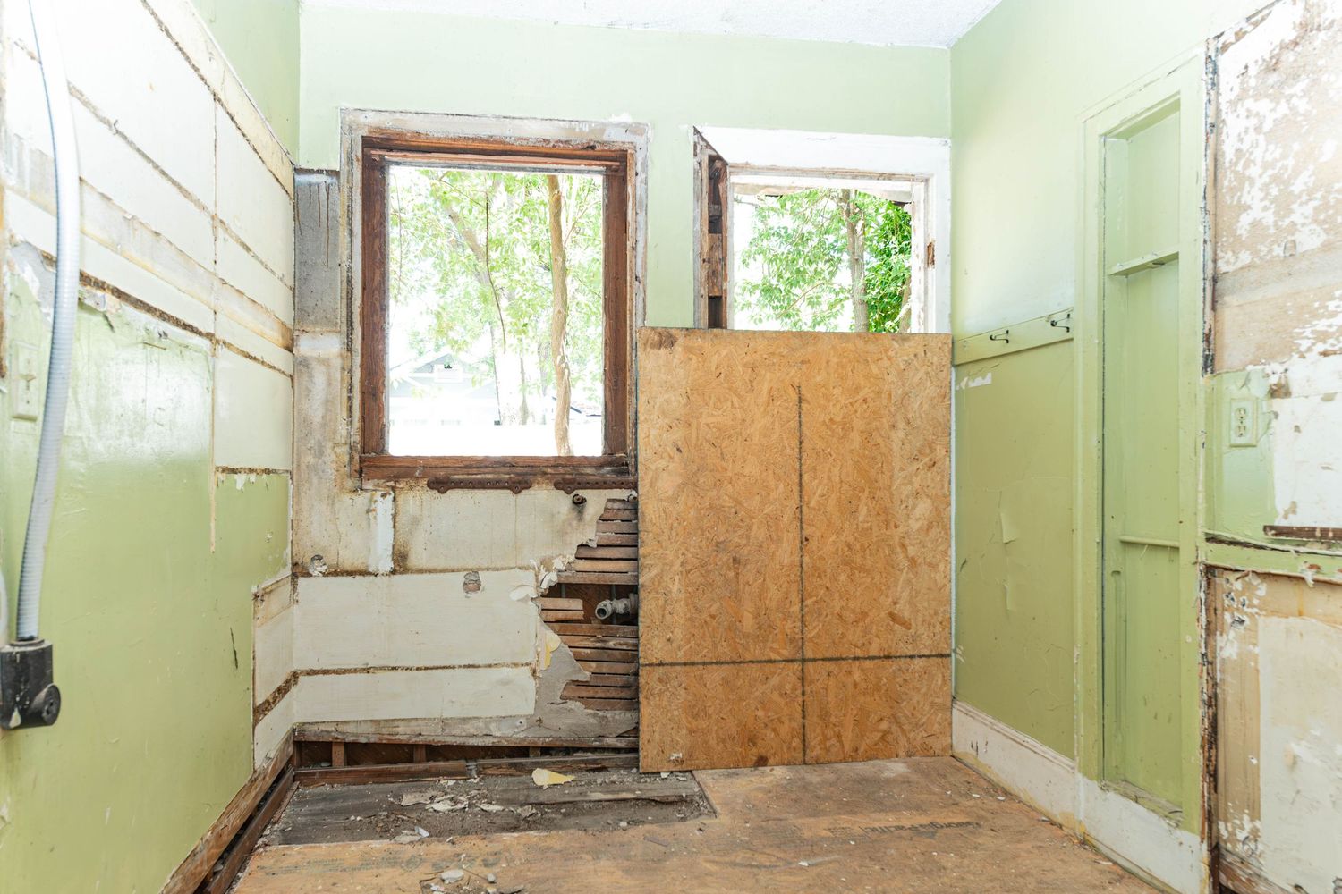 Dilapidated room with peeling green walls, damaged flooring, plywood barrier, and a window during home renovation.
