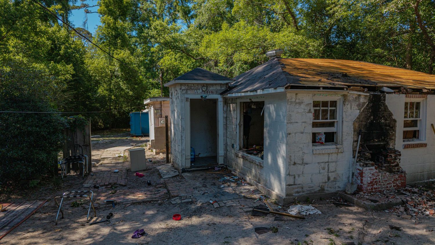 Abandoned dilapidated house with damaged walls, debris scattered on ground, surrounded by trees on a sunny day.