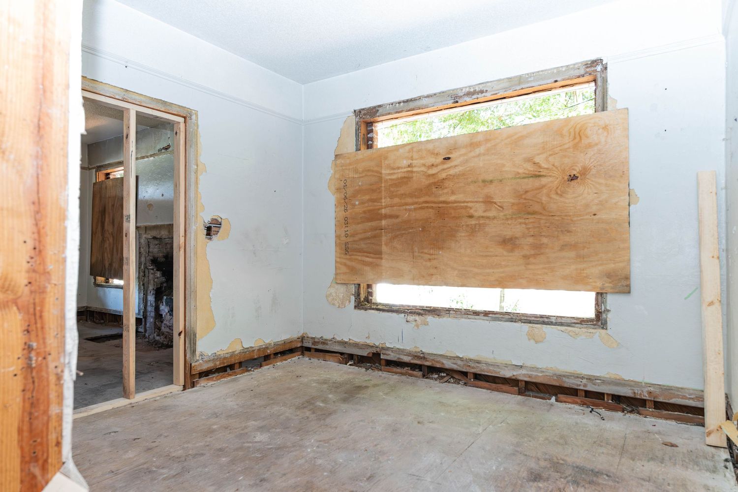 Unfinished room under renovation with plywood-covered window, exposed walls, and concrete floor.