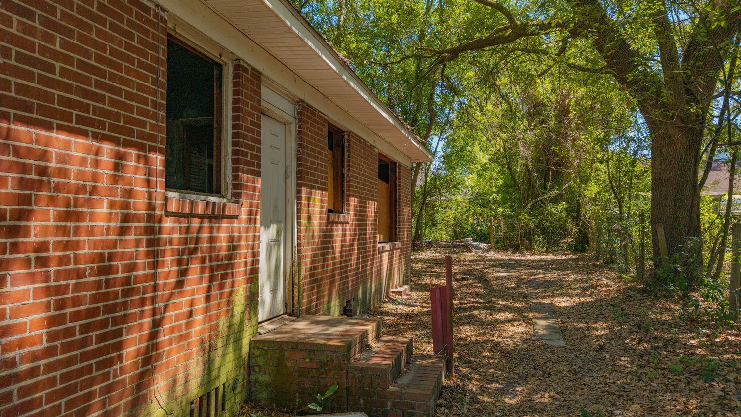 Brick ranch home exterior with wooded backyard, dirt path, and mature shade trees in a residential neighborhood.