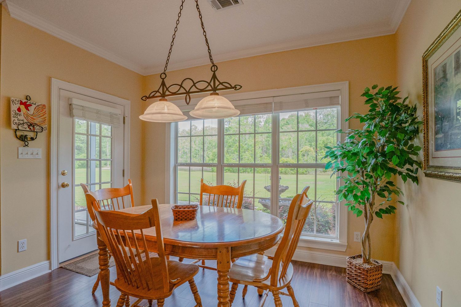 Bright dining area with oval wood table, chairs, large windows, hanging light fixture, and indoor plant on hardwood floors.