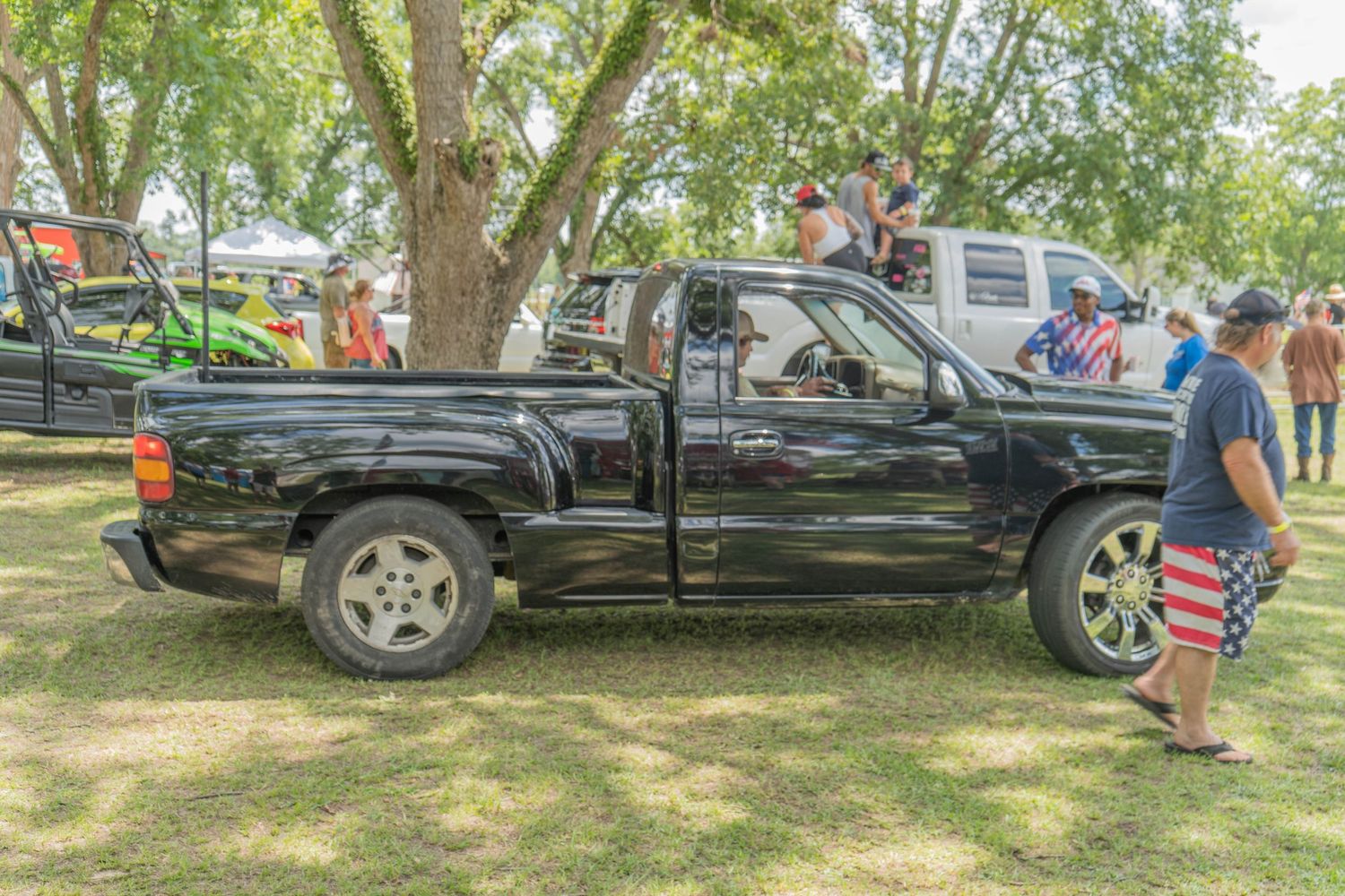 Black pickup truck parked on grass at an outdoor car show event with spectators and large trees in background.