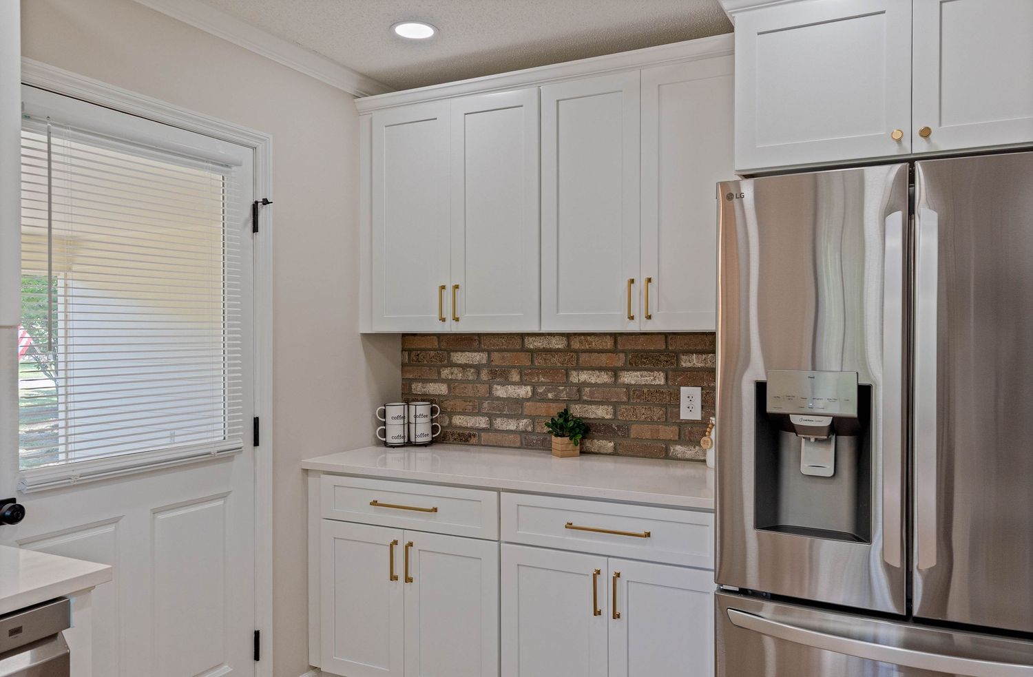 White kitchen cabinets with gold hardware, brick backsplash, and stainless steel refrigerator in a bright modern kitchen.