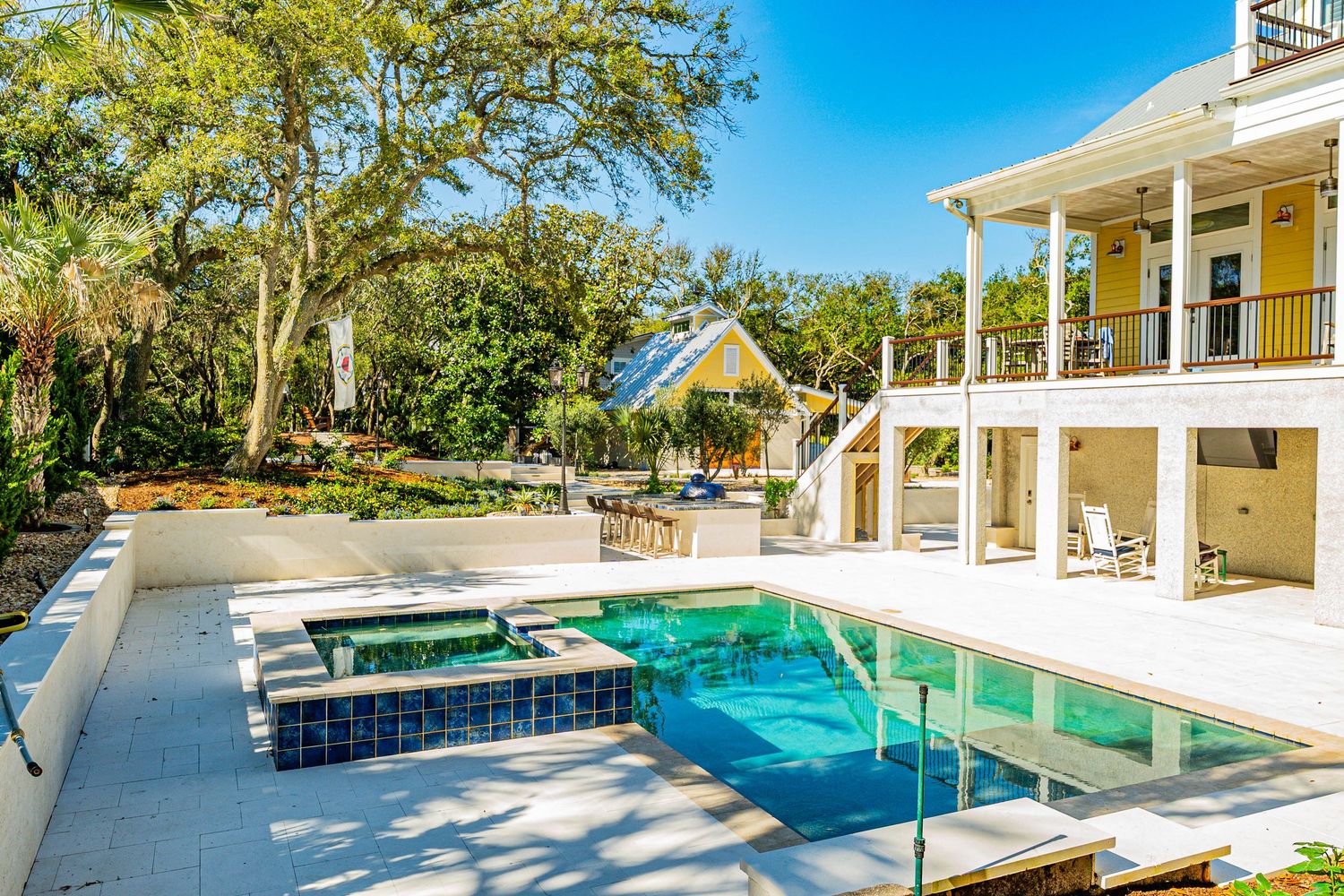 Luxury outdoor pool and spa with glass railings, tropical trees, and a yellow cottage near a two-story coastal home.