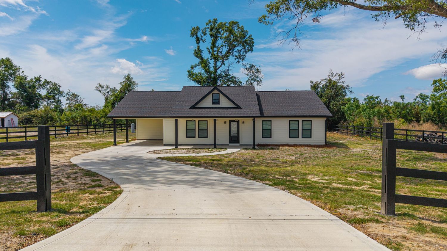 New ranch-style home with black fence, white trim, covered carport, and long concrete driveway surrounded by trees.