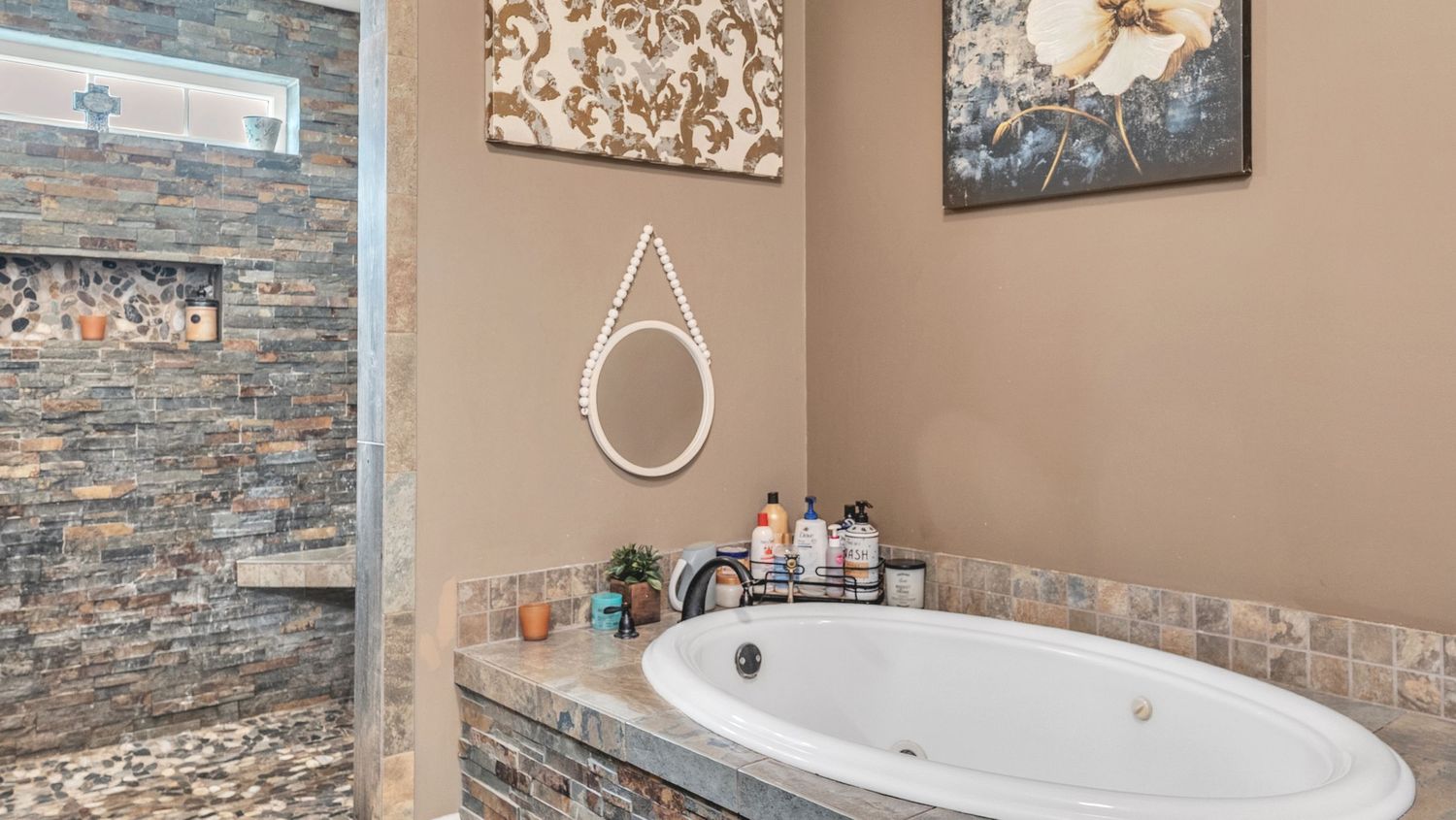 Luxurious bathroom featuring a stone-tiled soaking tub, round mirror, and decorative artwork on warm beige walls.