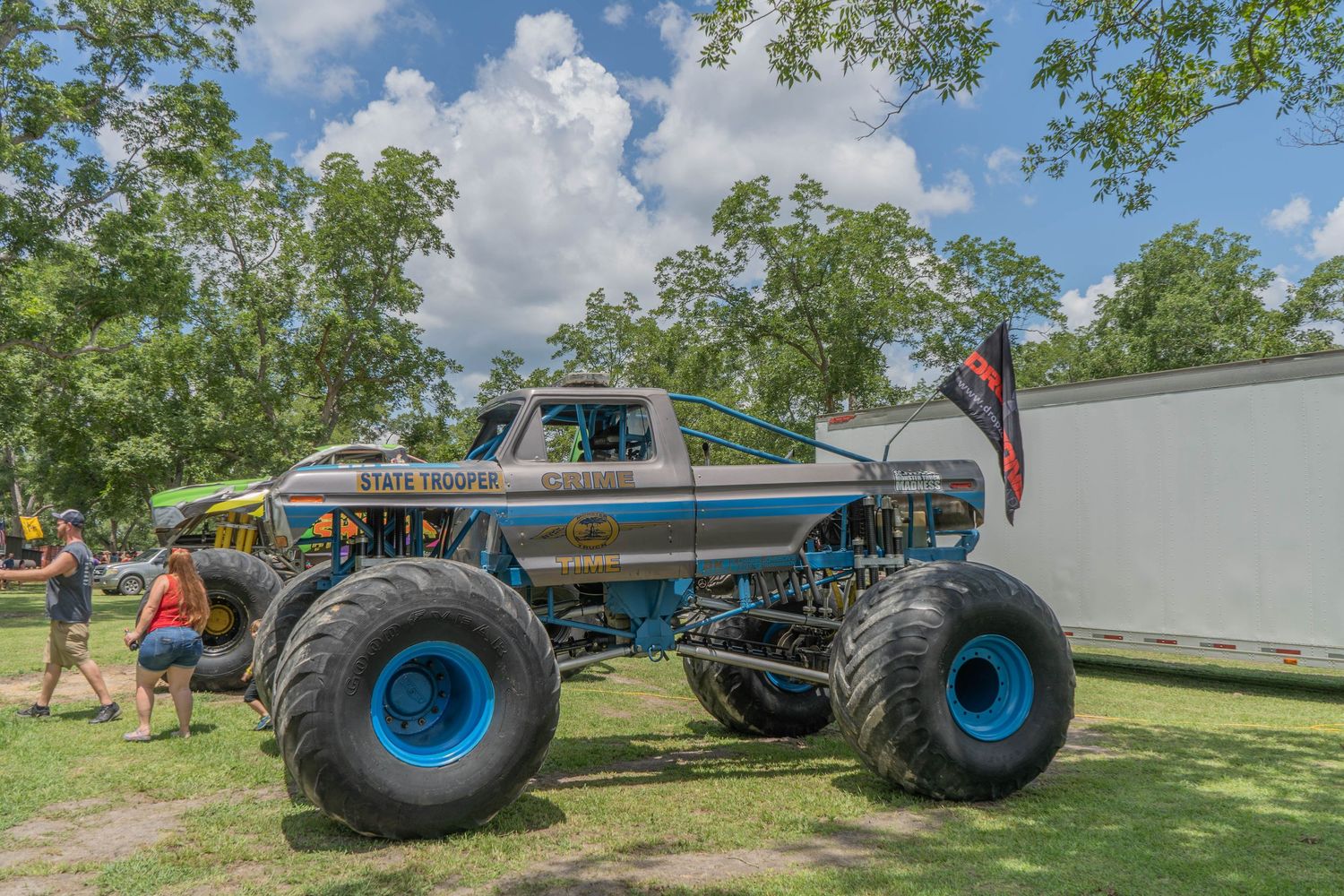A large monster truck with blue tires and a Confederate flag on display at an outdoor event surrounded by trees.