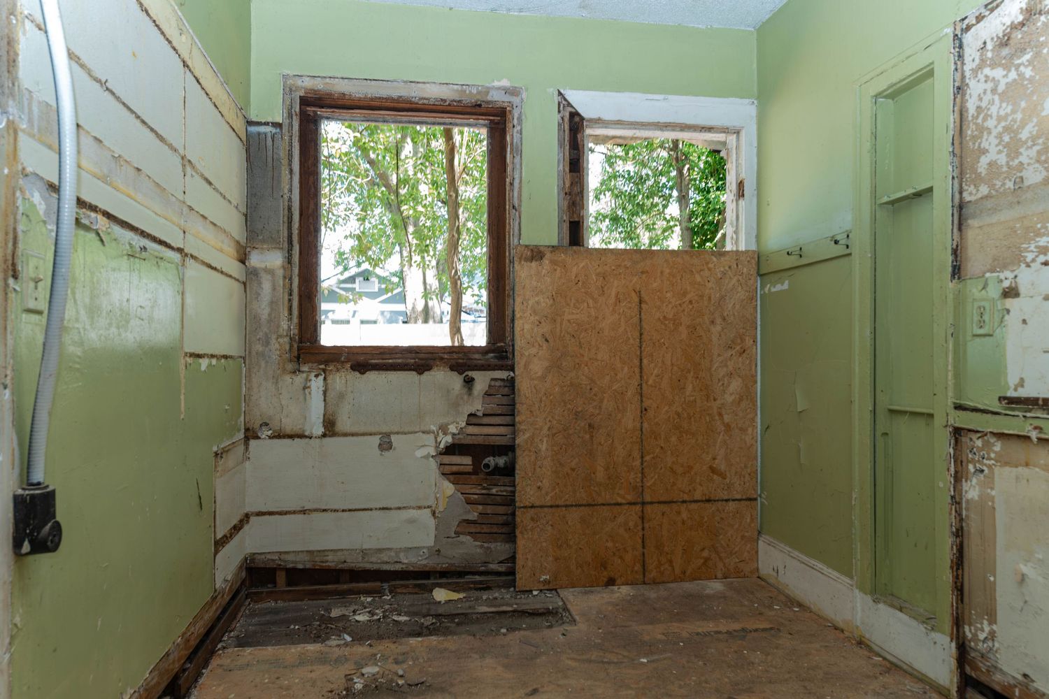 Abandoned room with peeling green walls, damaged flooring, and a boarded-up door with a window view of trees.