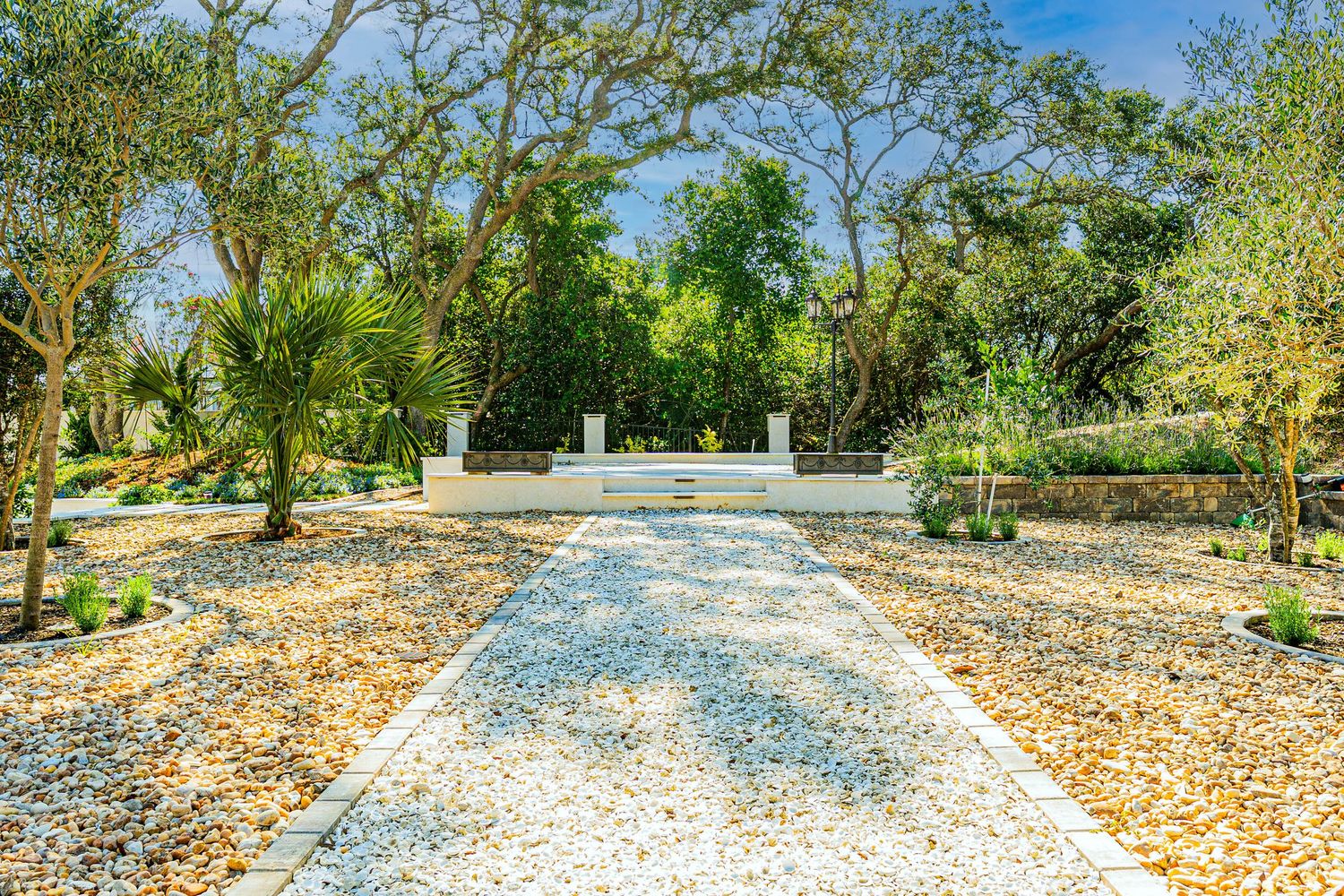 Serene garden pathway lined with gravel and lush trees leading to a white wall monument under a clear blue sky.