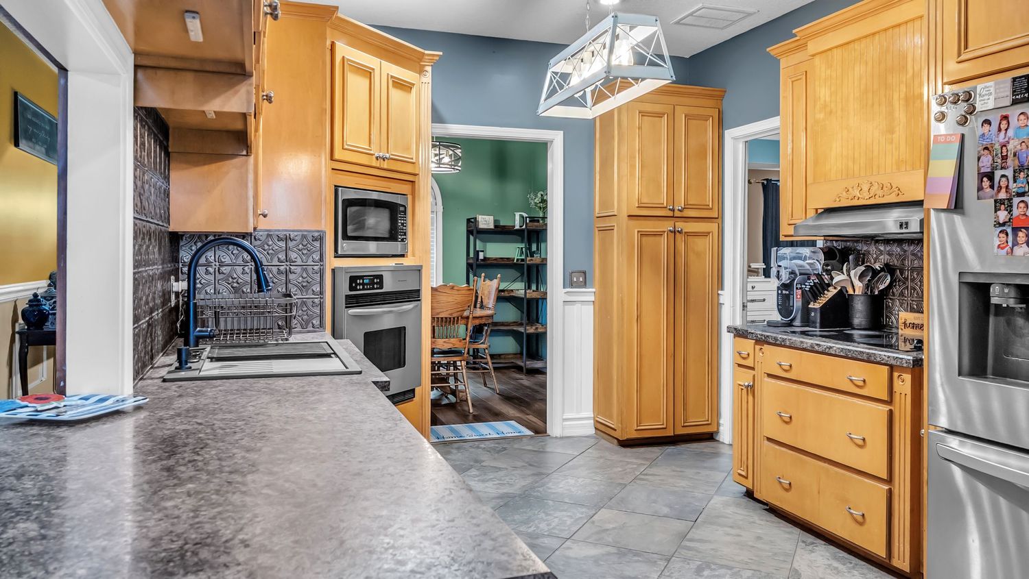 Modern kitchen with oak cabinets, stainless steel appliances, dark tile floors, and pendant lighting over a granite countertop.