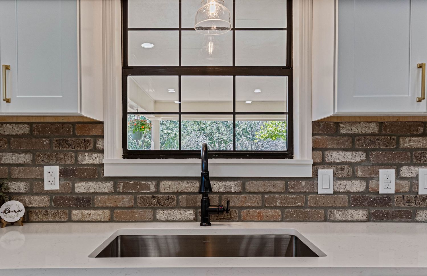 Modern kitchen sink with black faucet, brick backsplash, white cabinets, and large window with natural light.