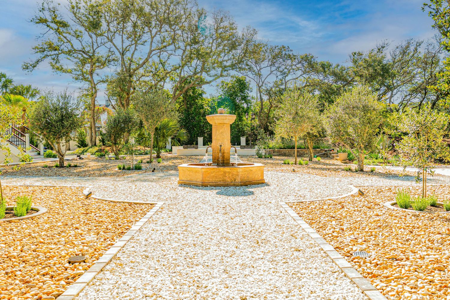 A serene garden path leads to a stone fountain surrounded by gravel and lush trees under a clear blue sky.