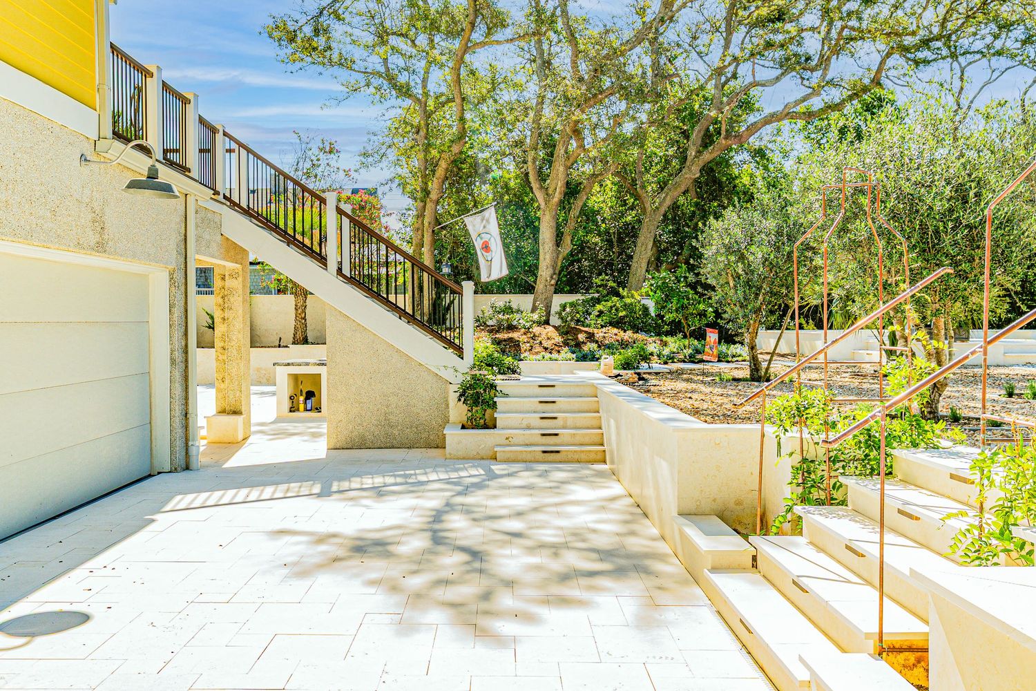 Modern villa exterior with white stone patio, elegant staircase, lush garden trees, and bright open-air terrace design.