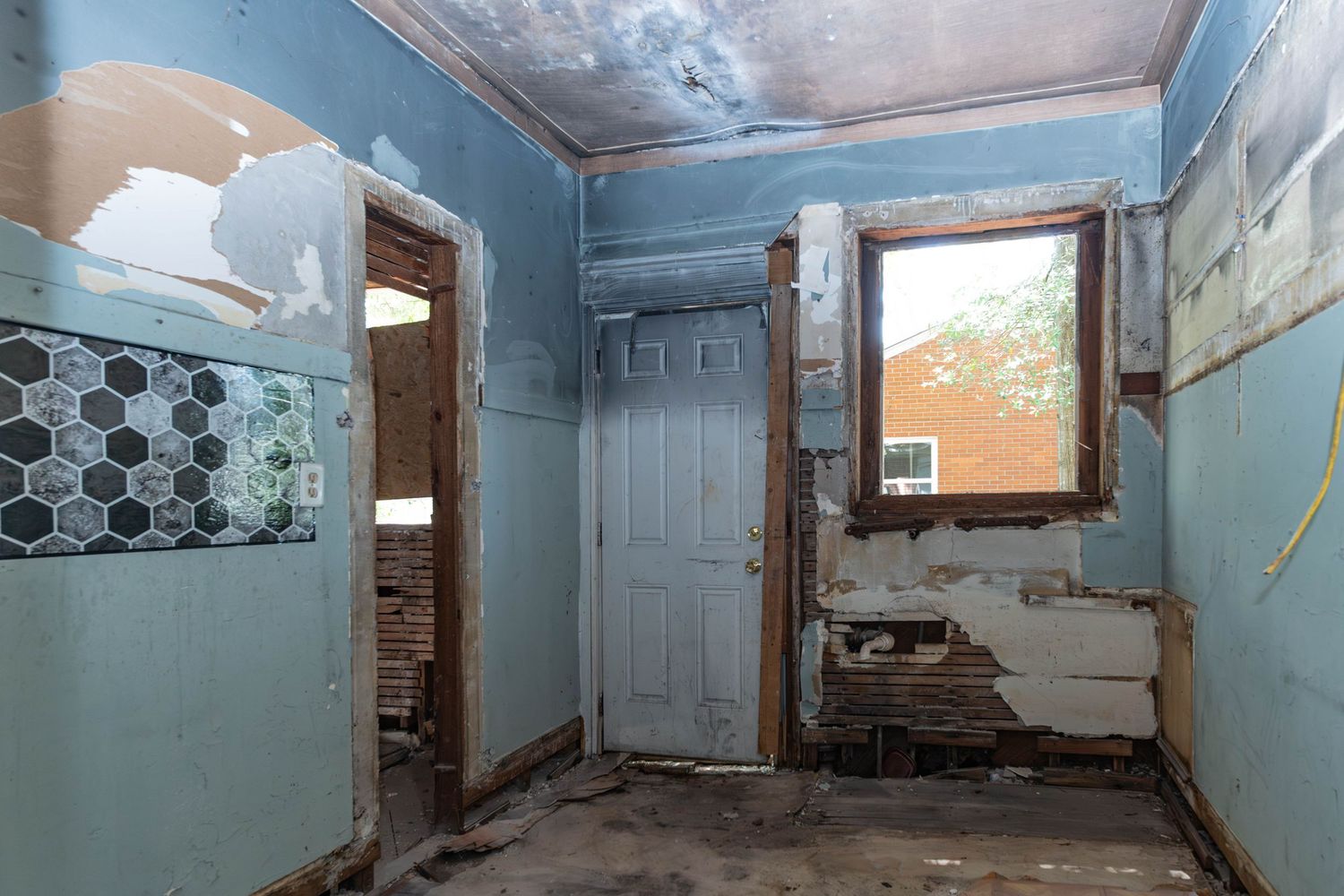Dilapidated room with peeling blue paint, exposed brick, deteriorating walls, and a window during abandoned home restoration.
