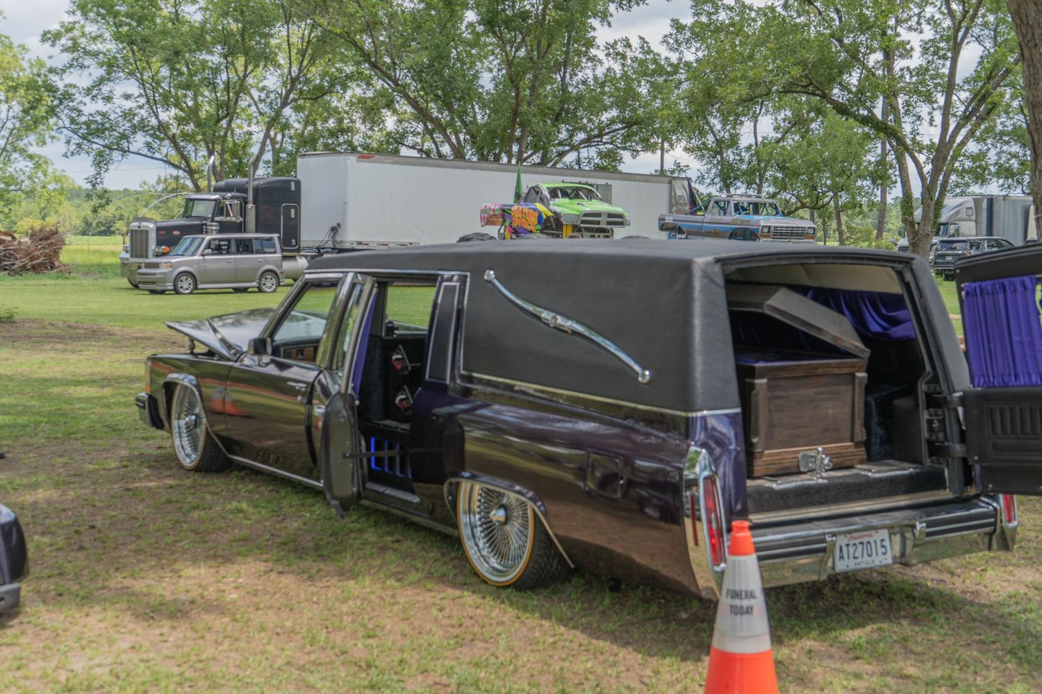 Black hearse with open casket compartment parked at outdoor event, orange traffic cone nearby.