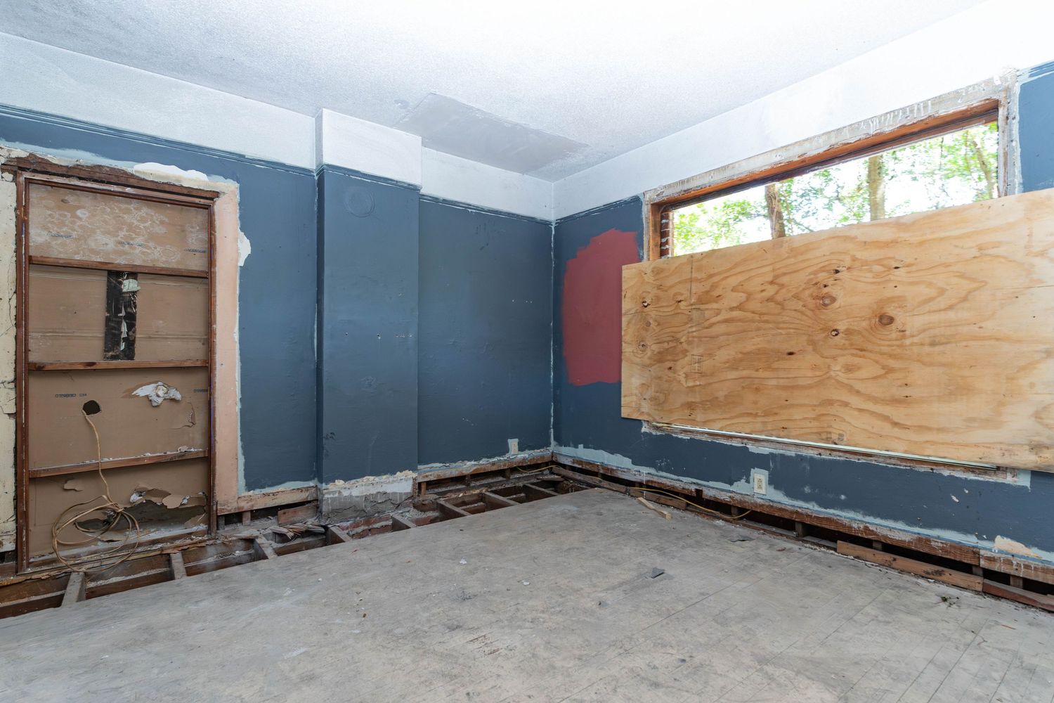 Gutted room with blue walls, boarded window, damaged flooring, and exposed door frames awaiting renovation.