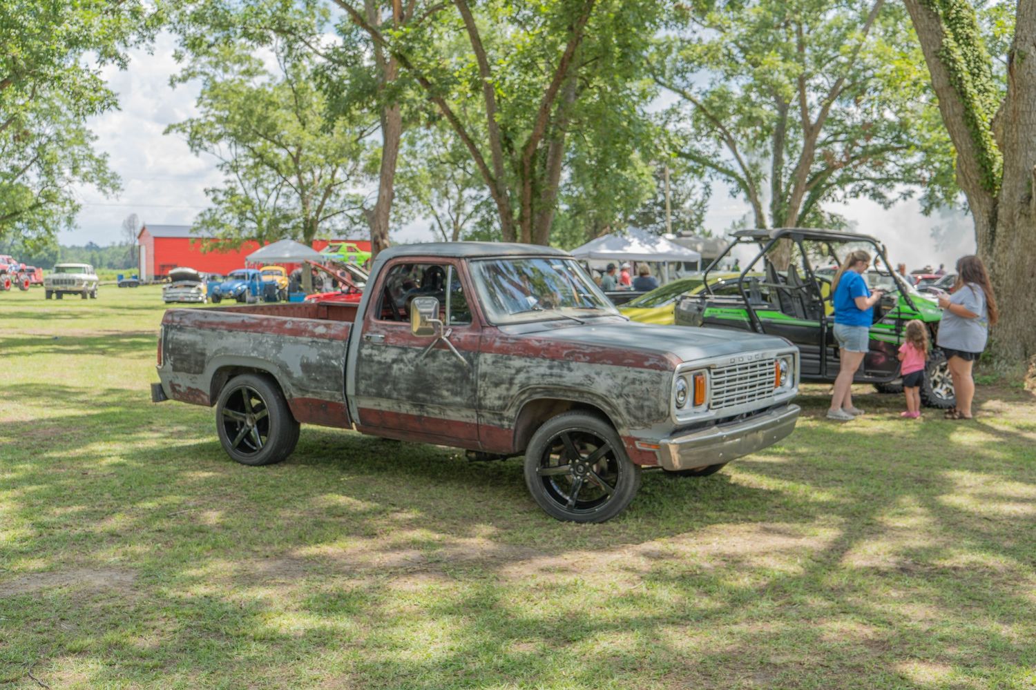 Rusty vintage pickup truck parked on grass at an outdoor car show with trees and spectators in background.