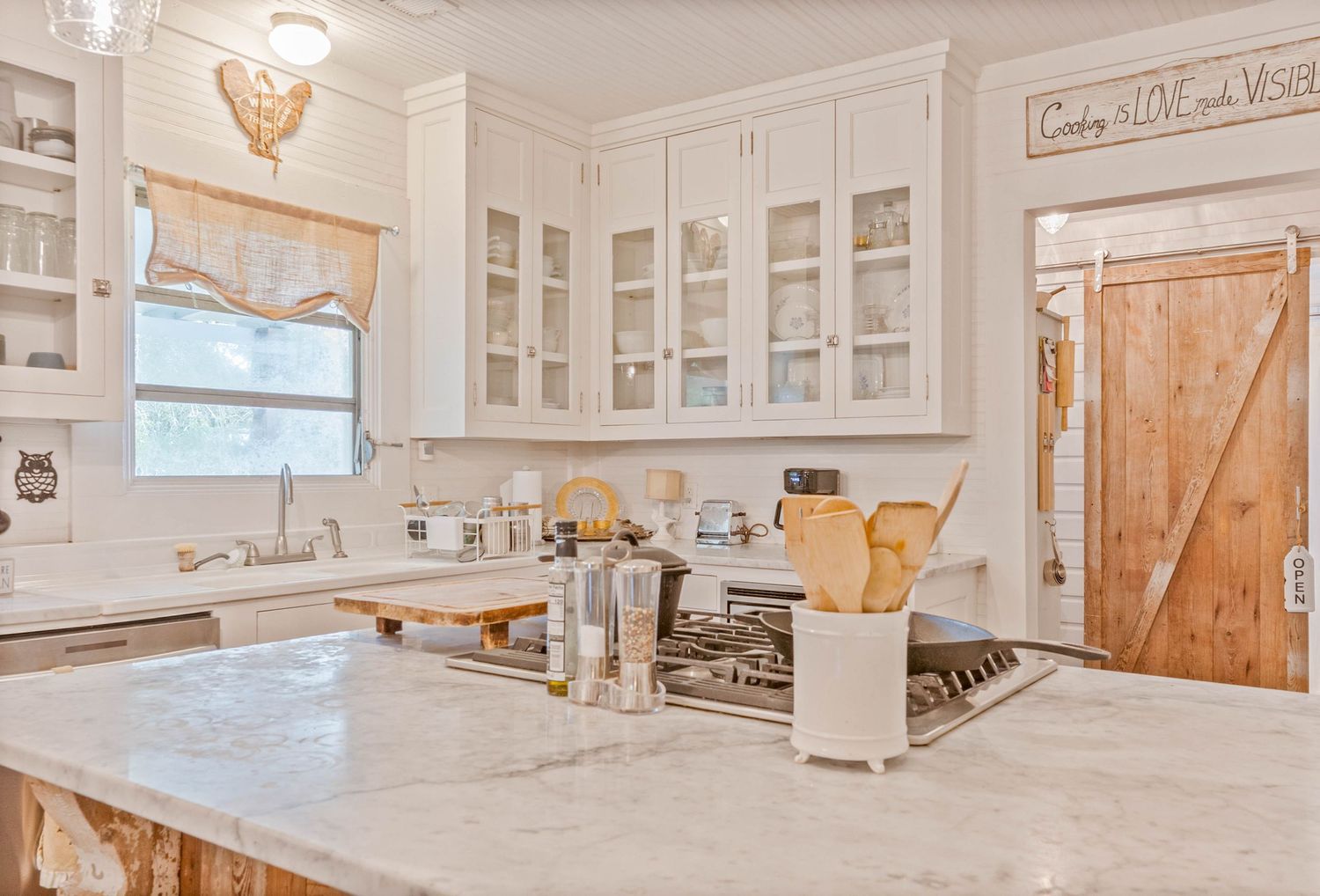 White farmhouse kitchen with marble island, glass-front cabinets, and rustic barn door accent.