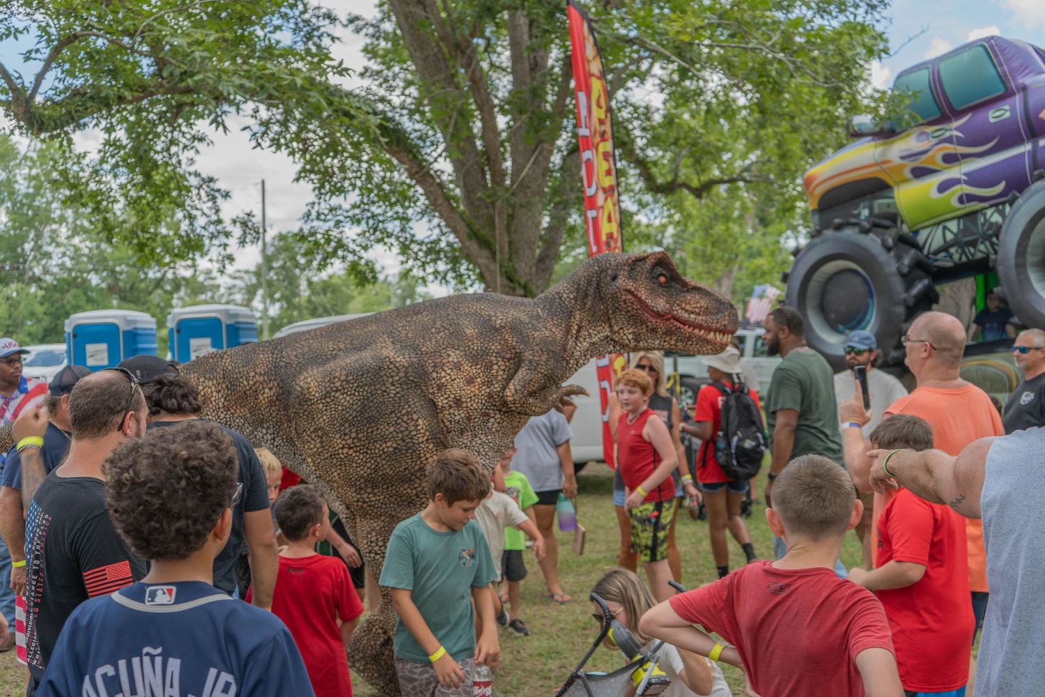 Crowd of people gathered around a realistic T-Rex dinosaur costume at an outdoor festival with a monster truck nearby.