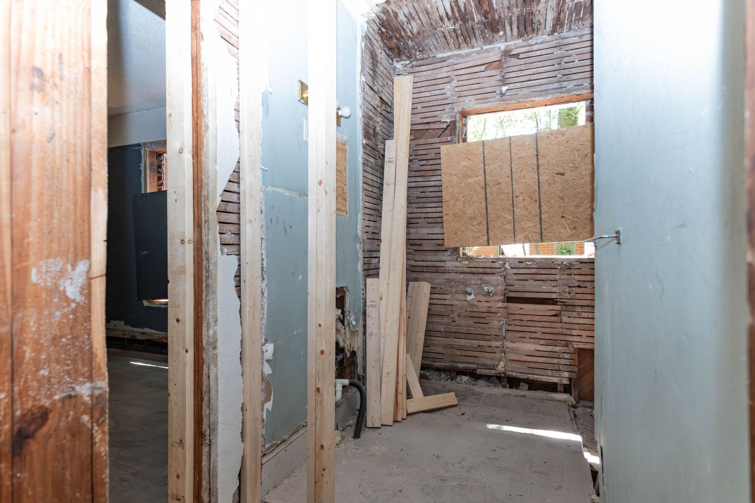 Narrow hallway under renovation with exposed wooden framing, drywall panels, and stacked building materials visible.