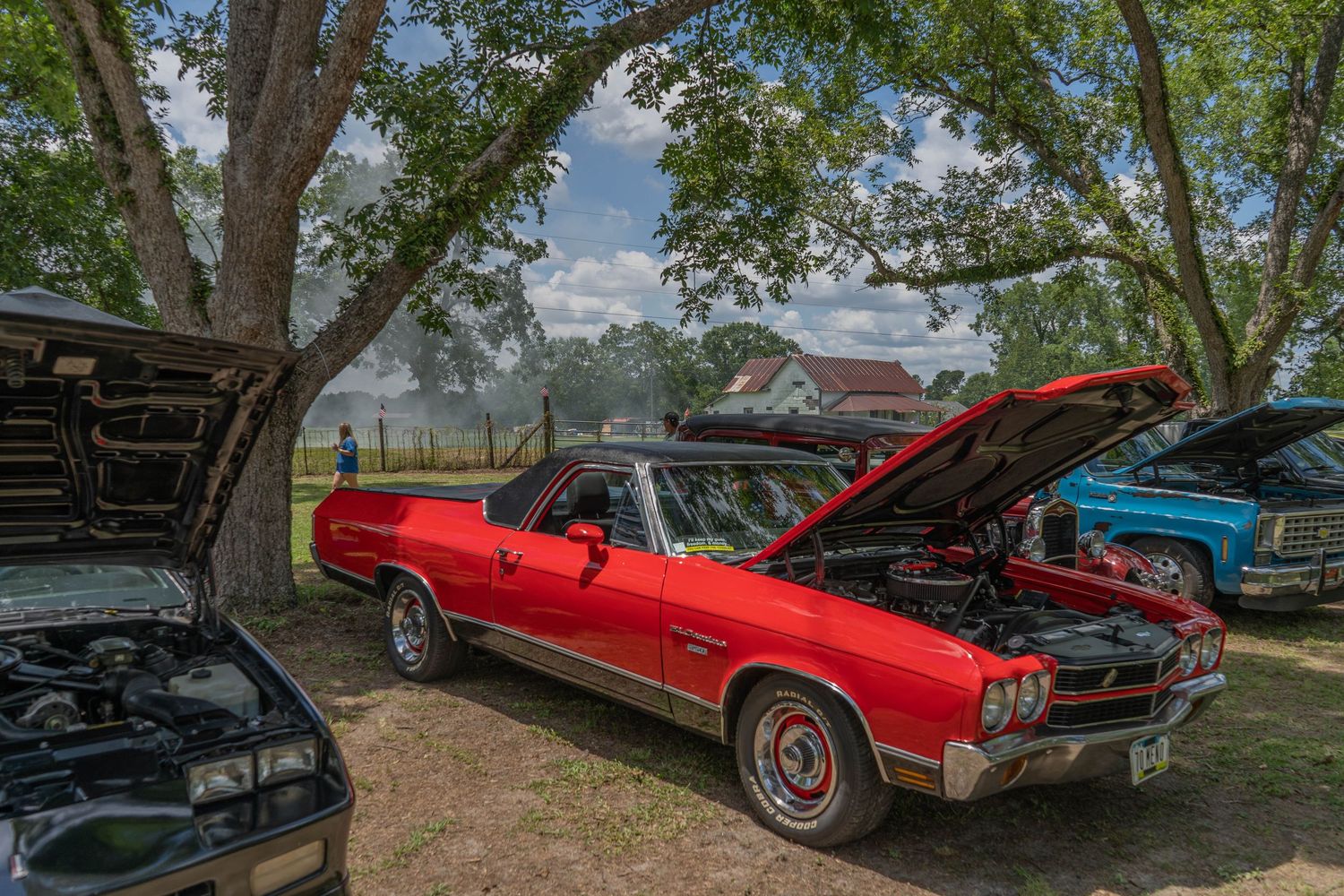 Red classic car with hood open at outdoor car show, parked under trees with other vintage vehicles on display.