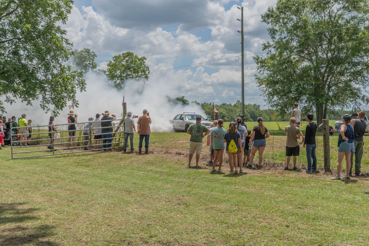 Crowd watches smoky car burnout at outdoor event on green field with trees and cloudy sky.