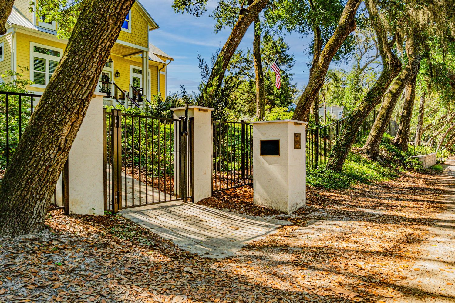 Black iron gate with white pillars leads to a tree-lined gravel driveway beside a yellow home.