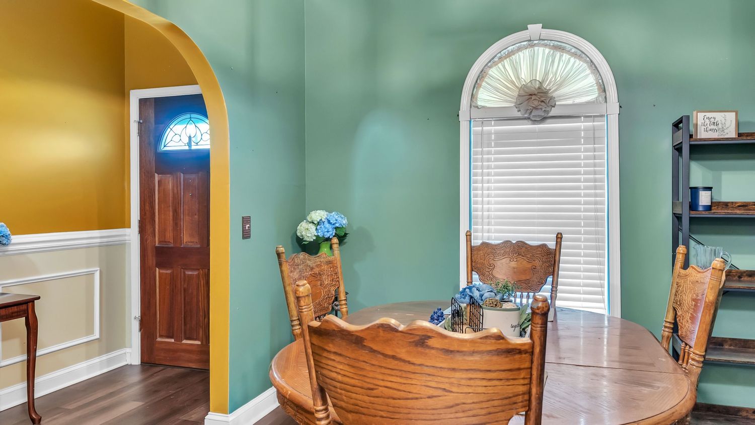 Cozy dining room with wooden table and chairs, arched windows, teal walls, and a view through an arched doorway to a foyer.