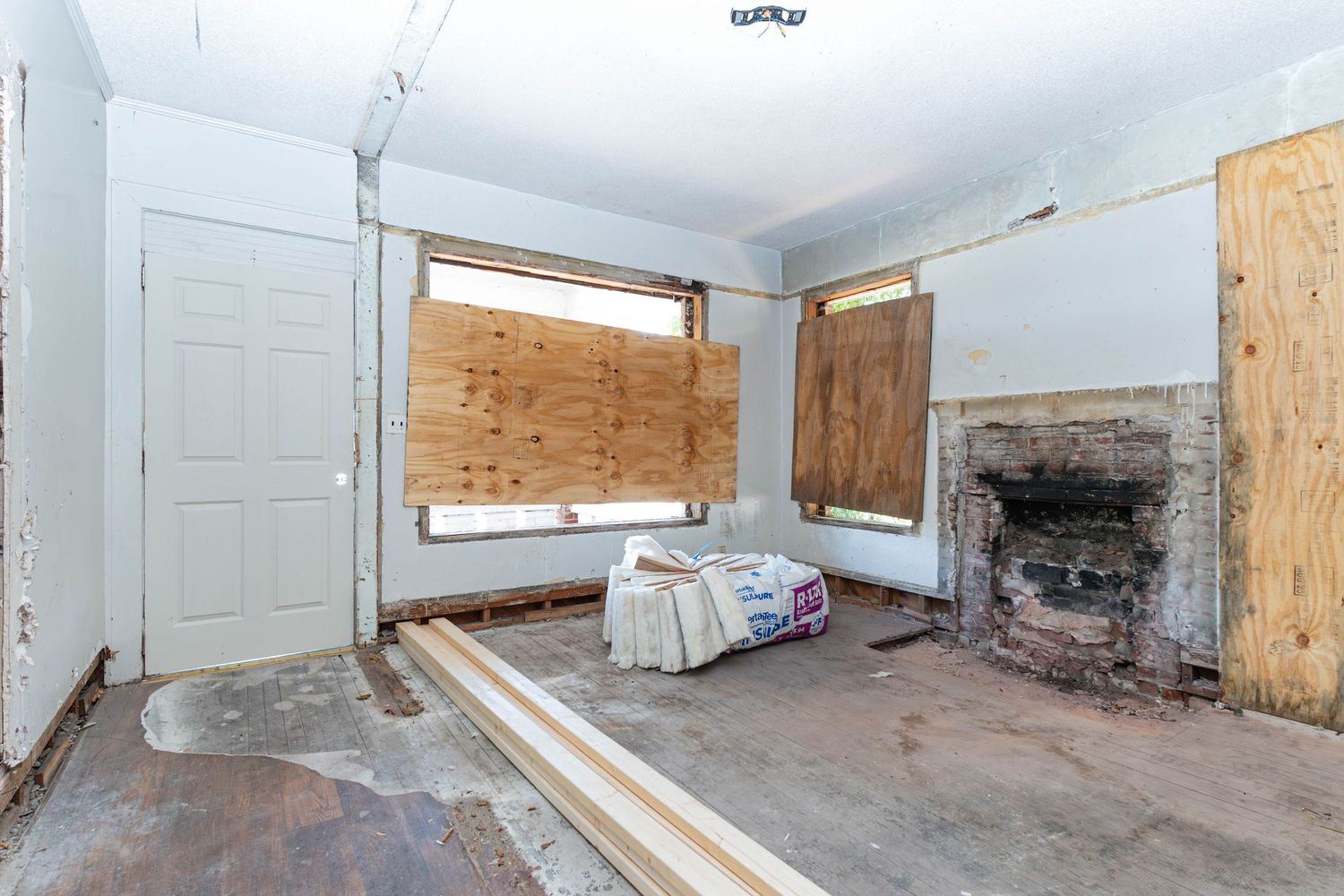 Empty room under renovation featuring boarded windows, brick fireplace, white door, and bare concrete floor.
