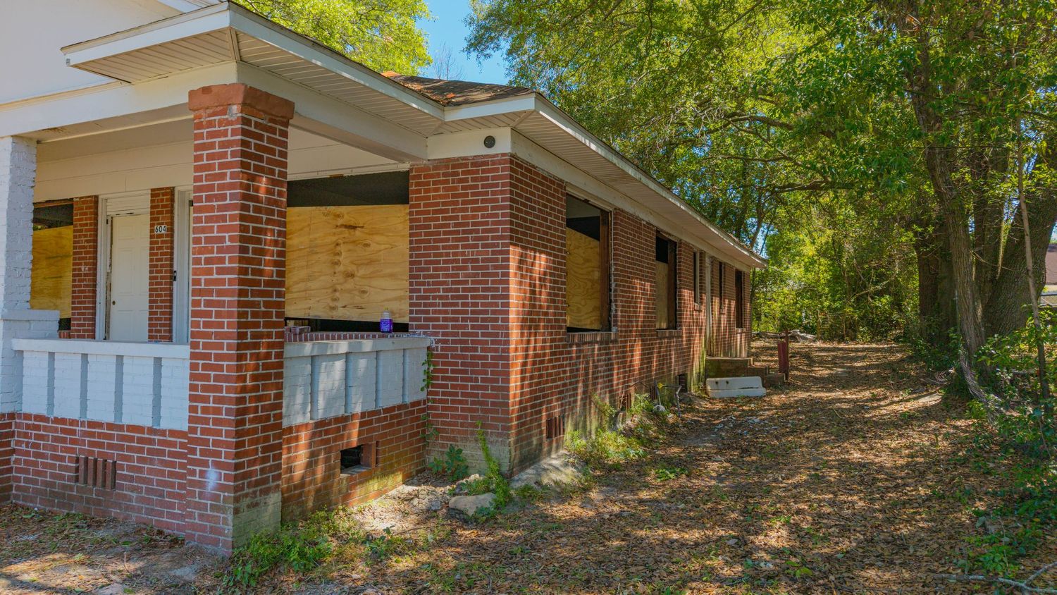 Abandoned brick house with boarded windows surrounded by overgrown trees and dry leaves in a neglected yard.
