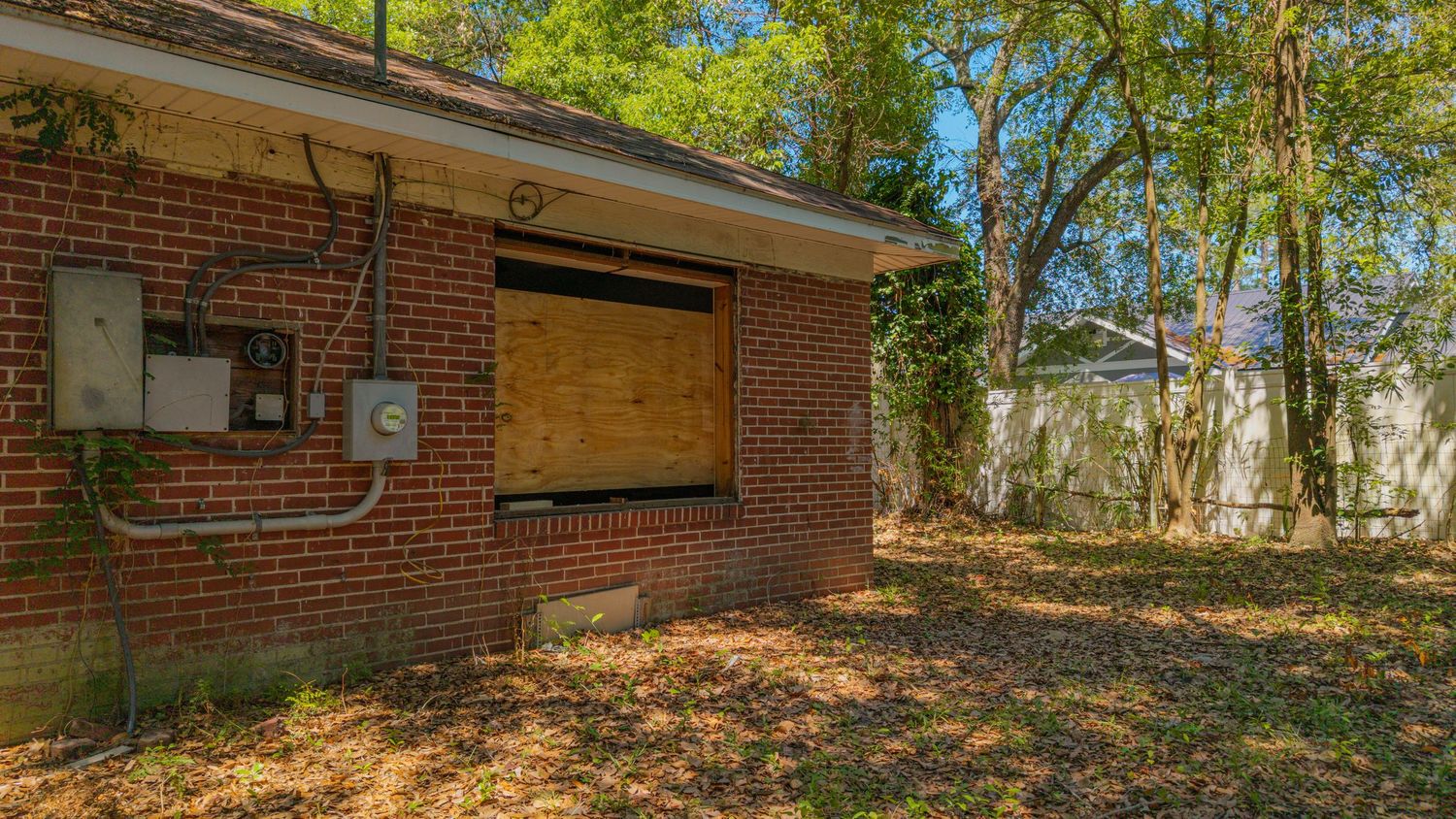 Abandoned brick building with boarded window, overgrown yard, and utility panel, surrounded by trees in a neglected property.