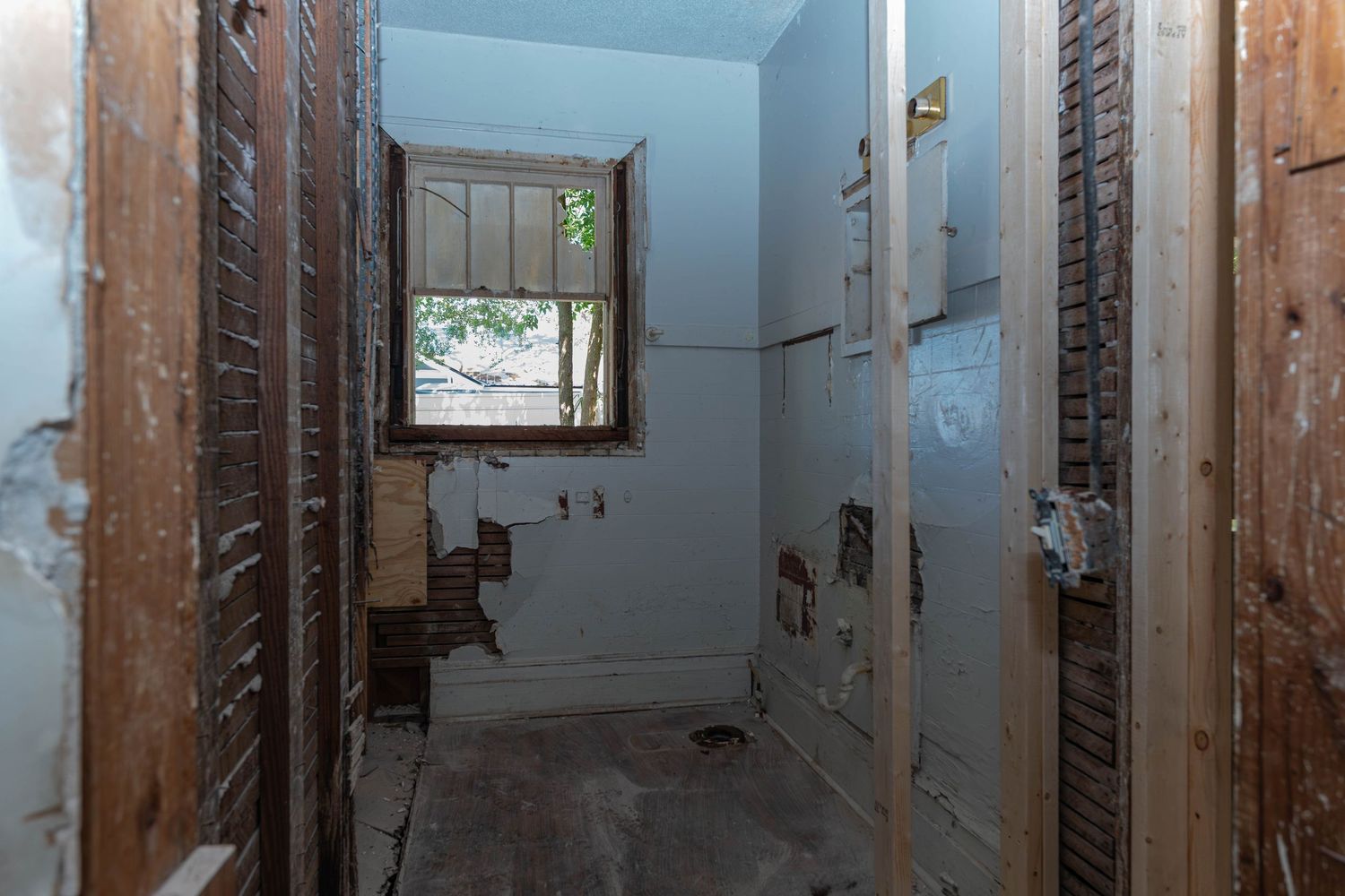 Gutted bathroom interior showing exposed walls, bare studs, and a single window during home renovation.