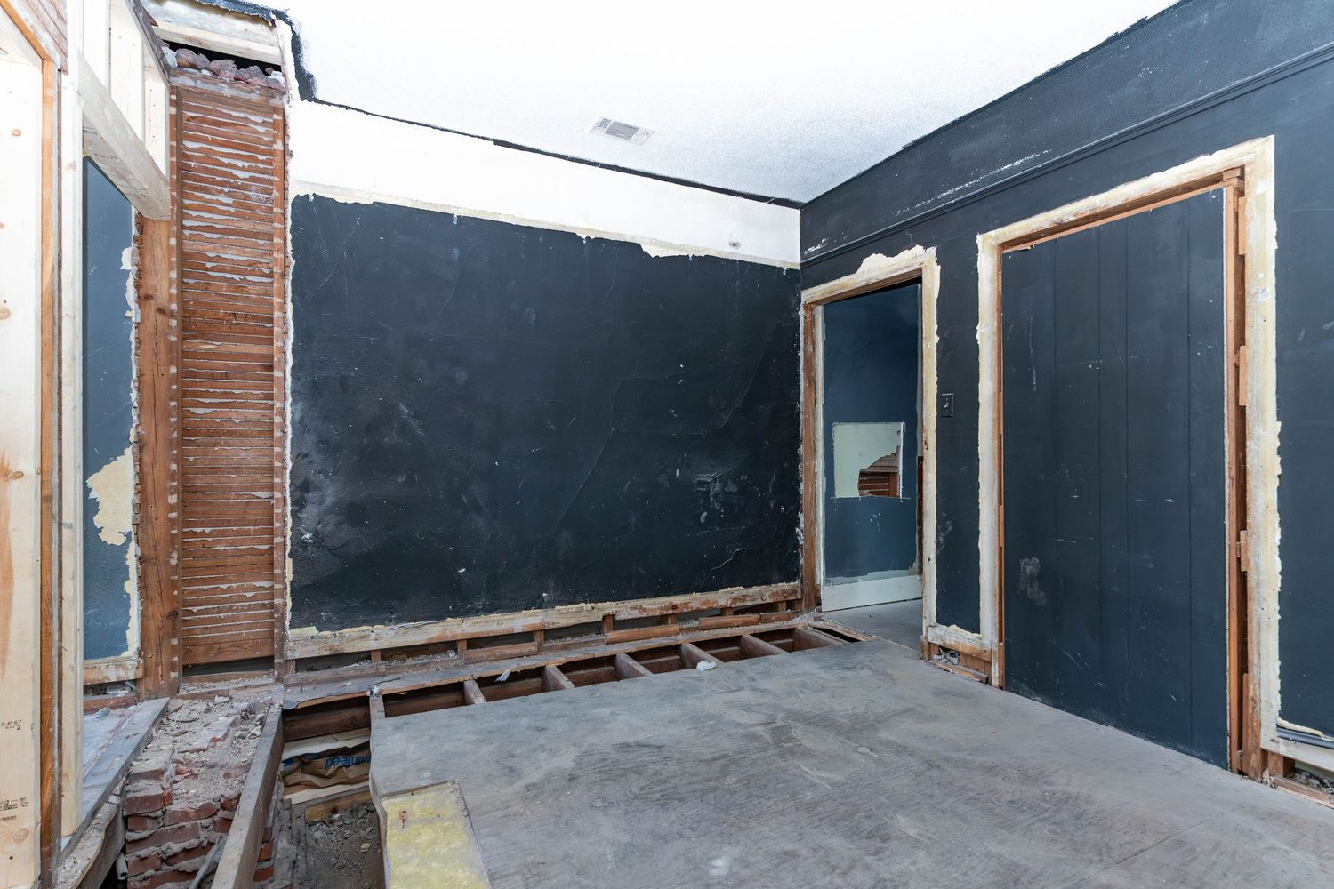 A gutted room under renovation with exposed wooden framing, dark walls, and bare concrete floors.