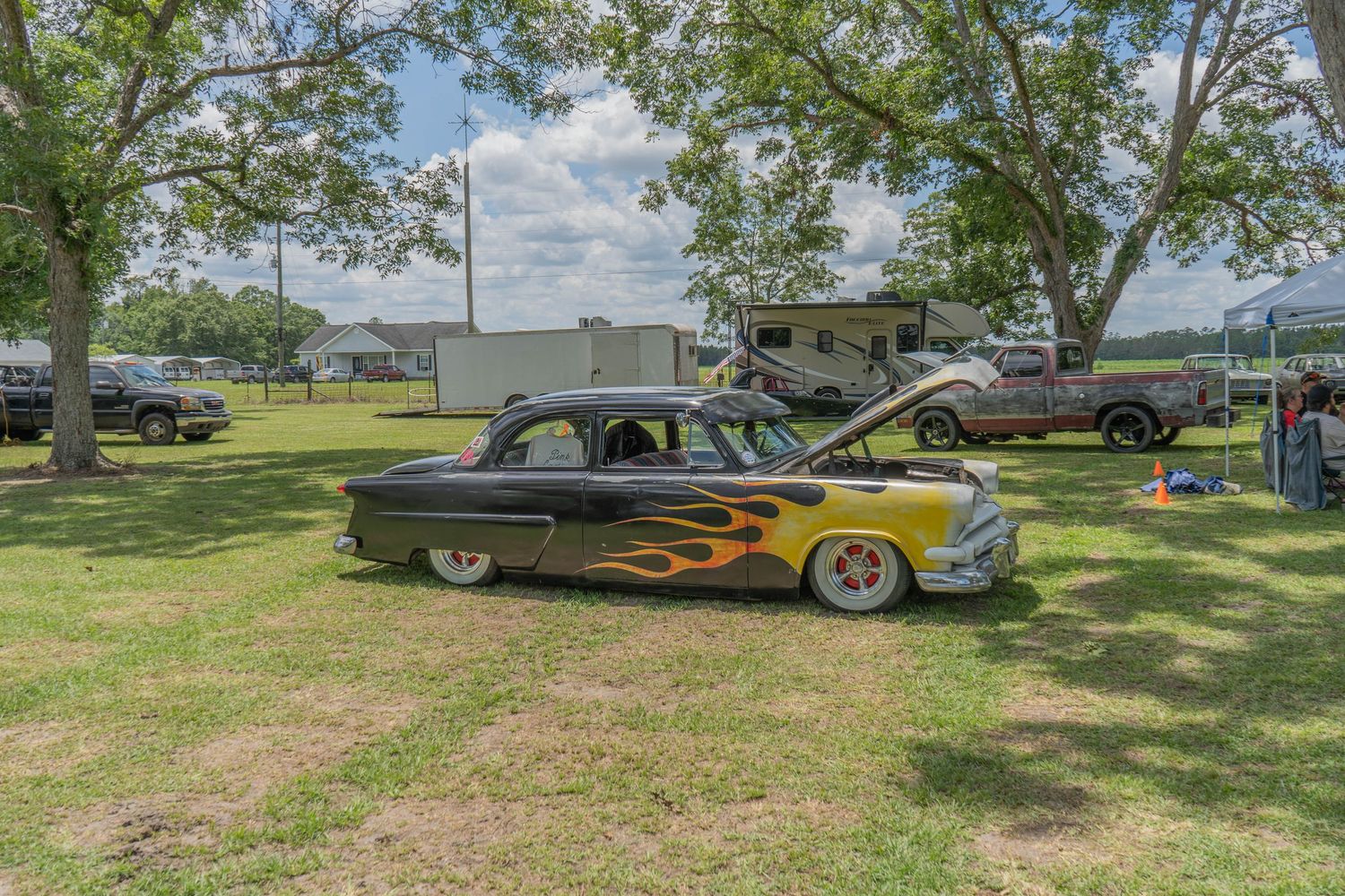 Classic black car with yellow flame paint job, hood open, displayed on grass at an outdoor car show.