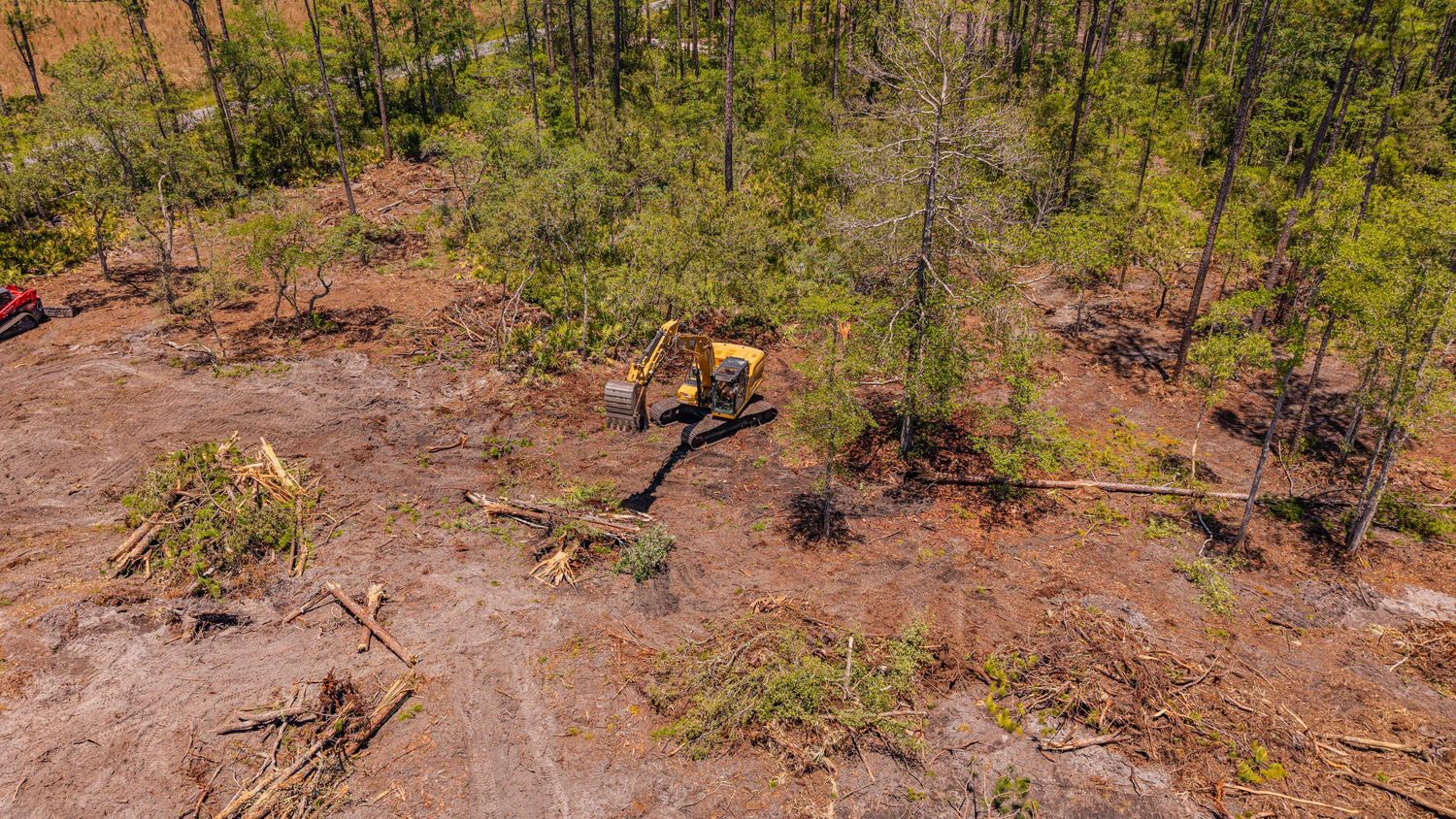 Aerial view of land clearing with excavator removing trees in a forested area with red clay soil.
