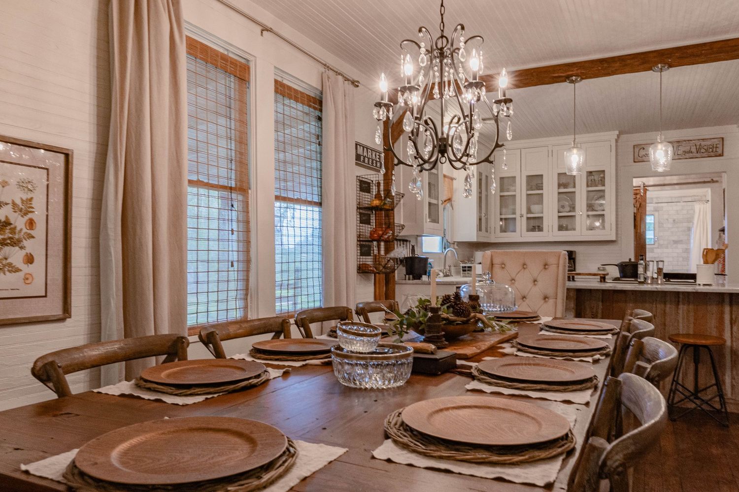 Rustic dining room with long wood table, chandelier, tall windows, and open kitchen view.