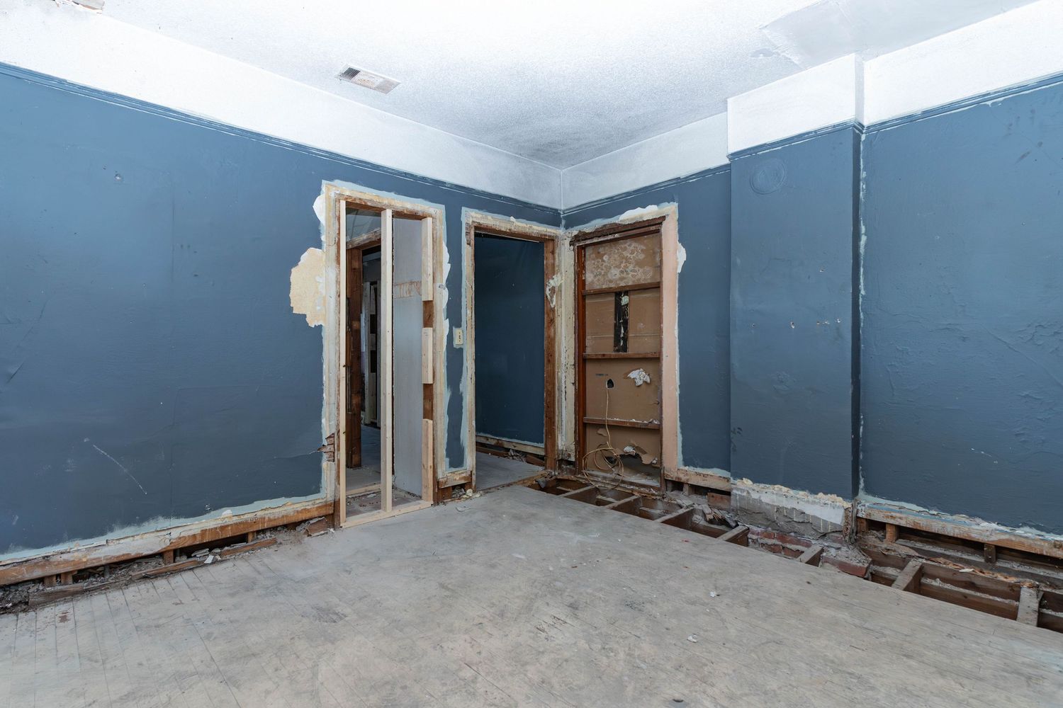 A room under renovation with blue walls, exposed door frames, and concrete floors in a gutted interior space.