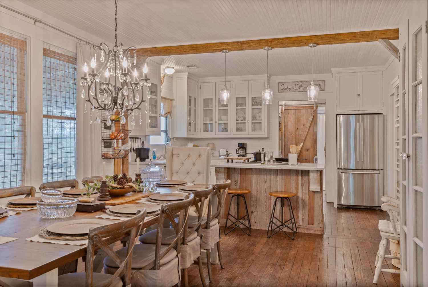 Farmhouse dining room with chandelier, wood table, upholstered chairs, white kitchen, barn door, and stainless steel fridge.