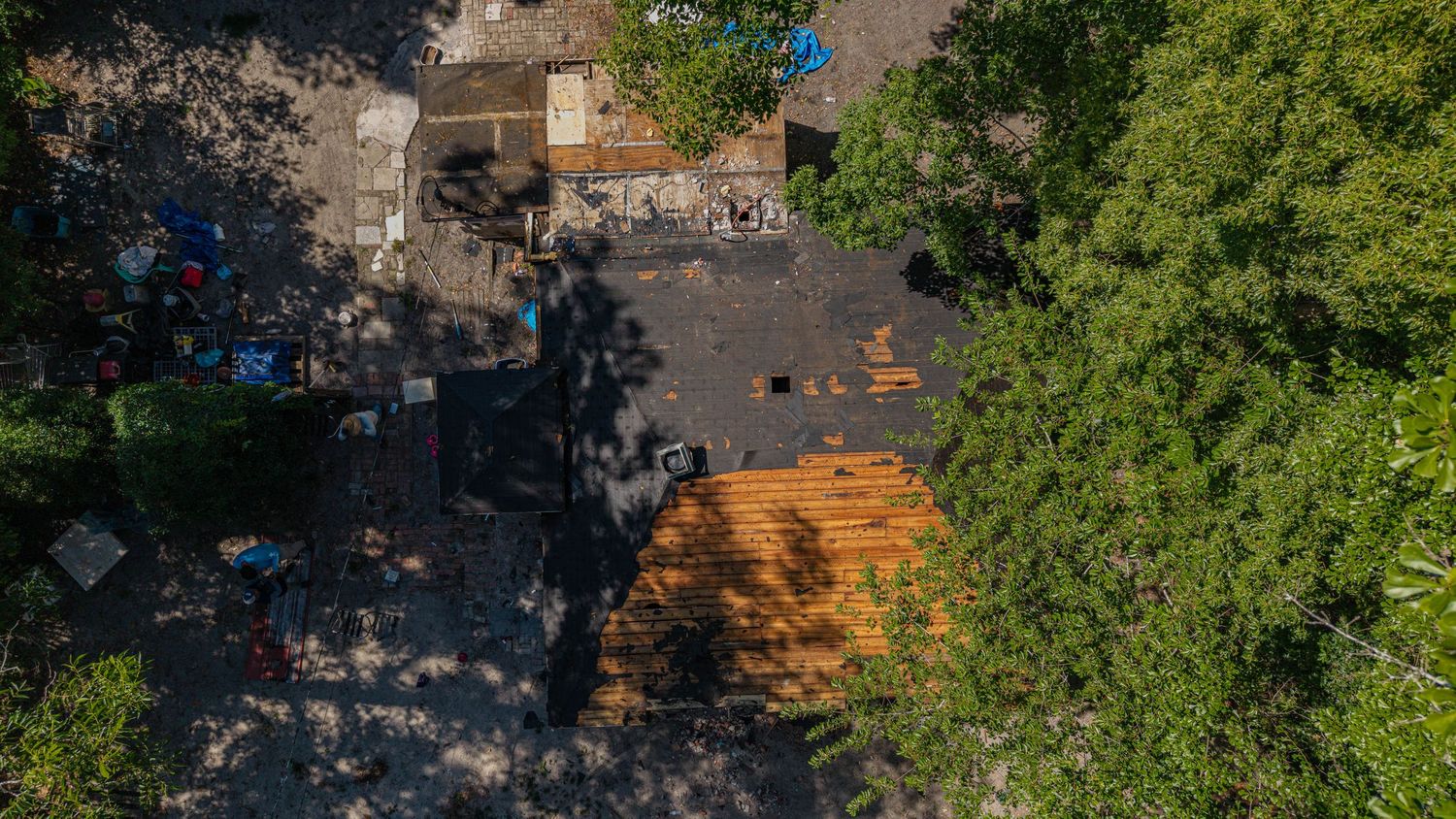 Aerial view of a residential rooftop undergoing repairs, surrounded by lush green trees and scattered debris.