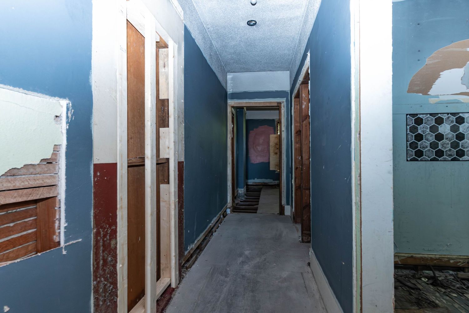 Narrow hallway under renovation with blue walls, exposed drywall, and multiple doorways in an older building.