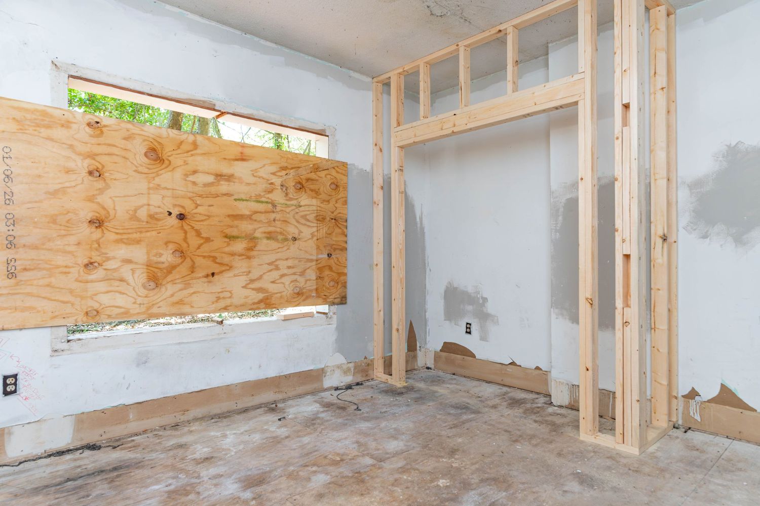 Interior room under renovation with boarded window, wood framing, and unfinished drywall walls.