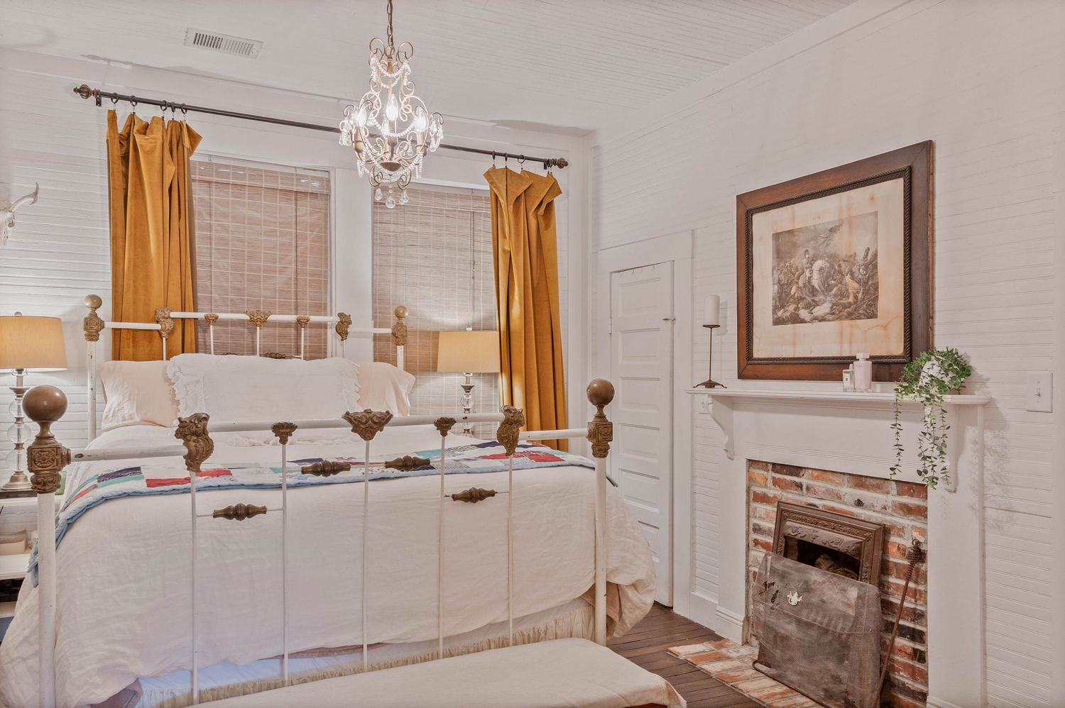 Elegant bedroom with white iron bed, gold curtains, crystal chandelier, and brick fireplace in a charming vintage-style room.