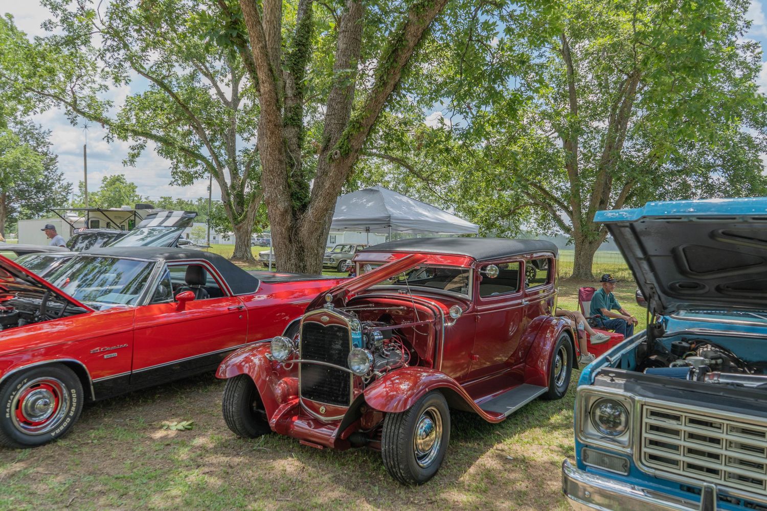 Vintage red hot rod at an outdoor classic car show with trees and other classic vehicles in the background.