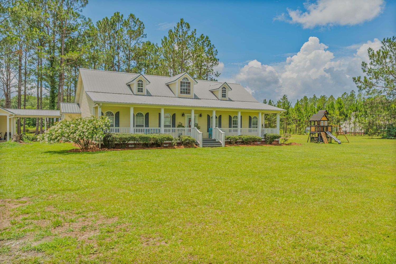 White farmhouse with wraparound porch surrounded by lush green lawn and tall pine trees under a blue sky.
