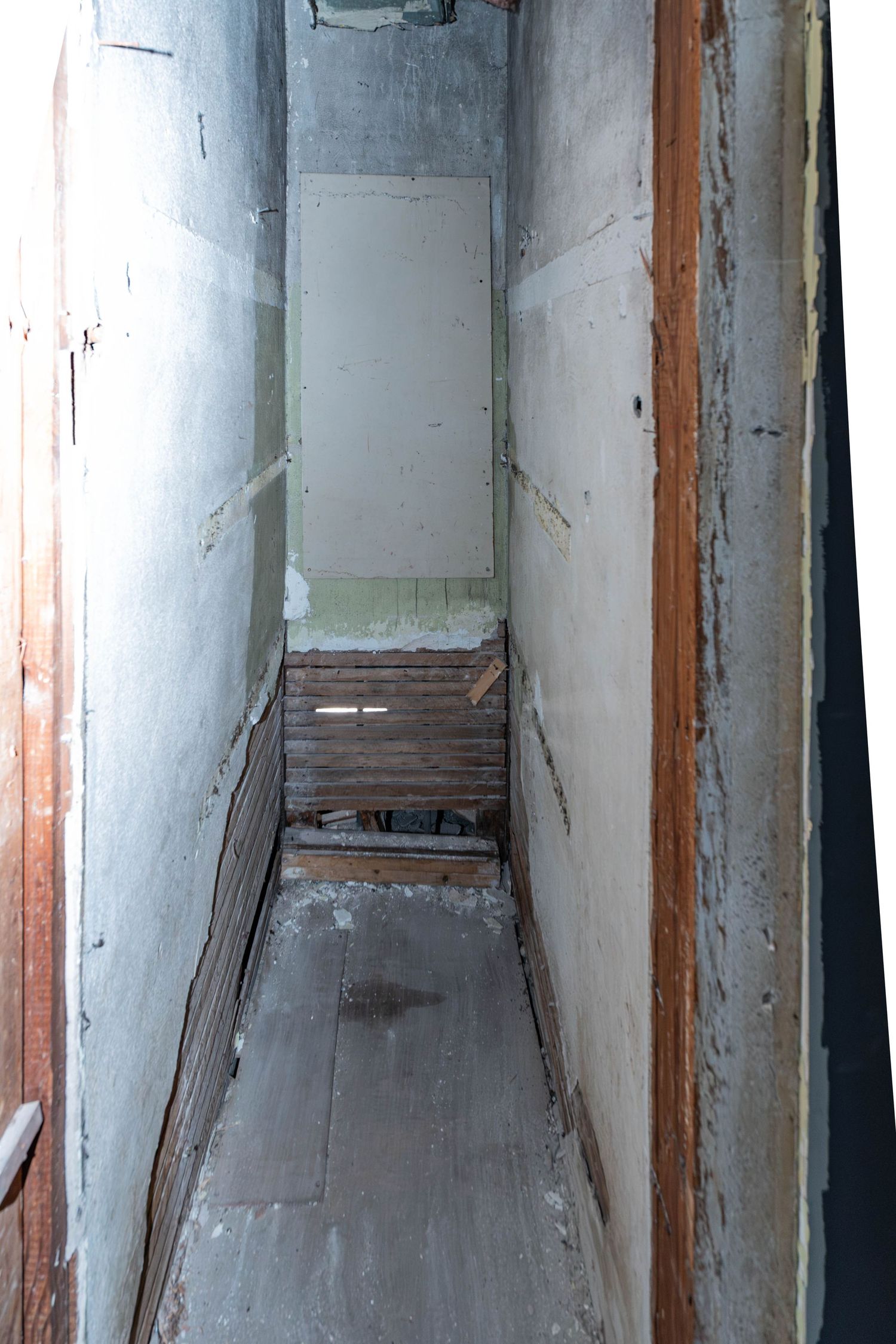 Narrow deteriorating hallway with damaged flooring, peeling walls, and broken wooden boards in an abandoned building.