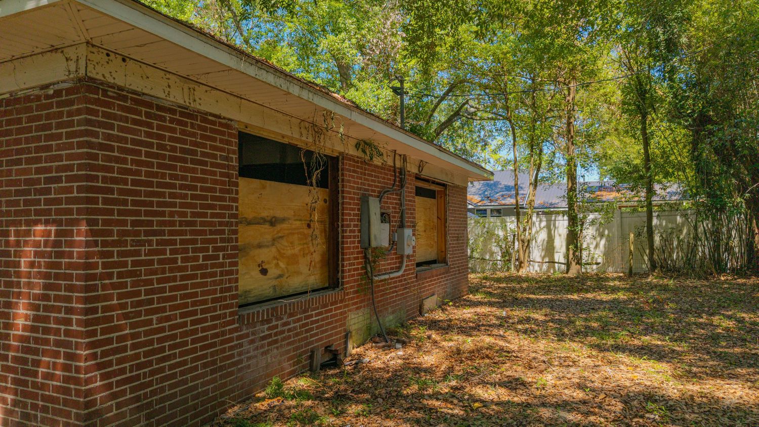 Abandoned brick ranch house with boarded windows surrounded by overgrown trees and neglected yard.