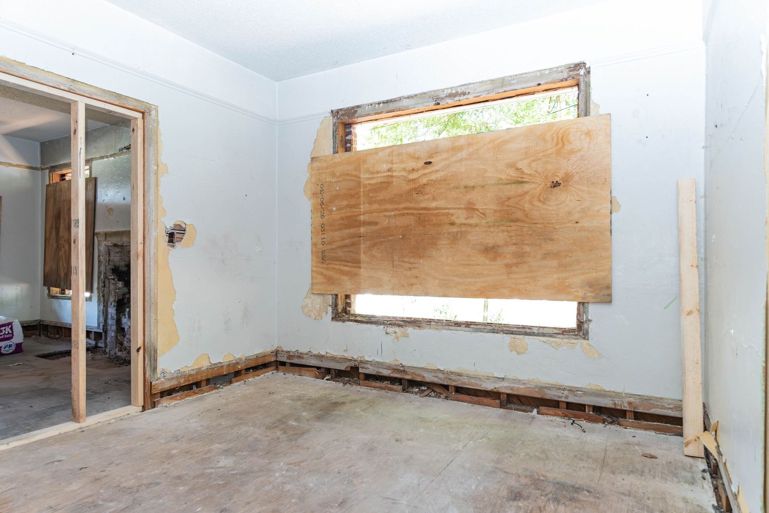 Room under renovation with plywood-covered window, exposed drywall, and bare concrete floor in a gutted interior space.