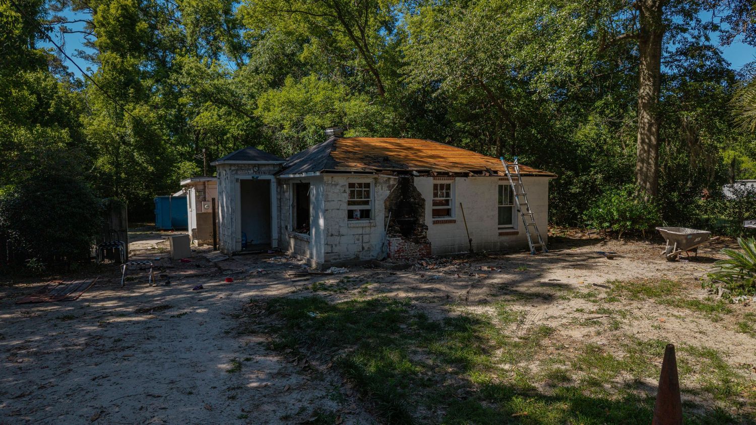 Abandoned single-story house with deteriorating roof surrounded by overgrown trees and sandy driveway in a rural setting.